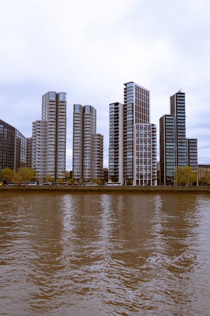 A panoramic view of a modern city skyline with high-rise office buildings and residential towers, situated along a riverbank. In the foreground, there is a calm body of water reflecting the buildings 