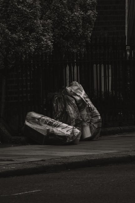 Two large black bin bags filled with waste materials rest on the pavement next to a sidewalk; the bags appear to be made of plastic with glossy finishes, crumpled and slightly torn, with some printed 