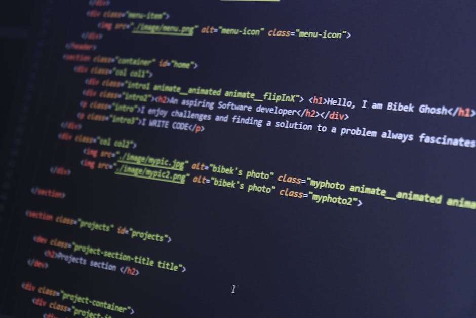 A person operating a silver MacBook laptop displaying a code editor with multiple lines of multicoloured programming code on a dark background. The individual's hands are visible, with the right hand